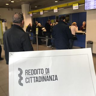 Persone in coda, mentre un cartello in primo piano riporta la scritta &quot;reddito di cittadinanza&quot;