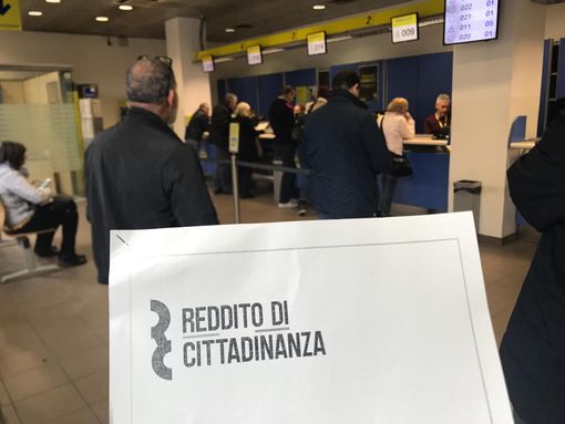 Persone in coda, mentre un cartello in primo piano riporta la scritta &quot;reddito di cittadinanza&quot;